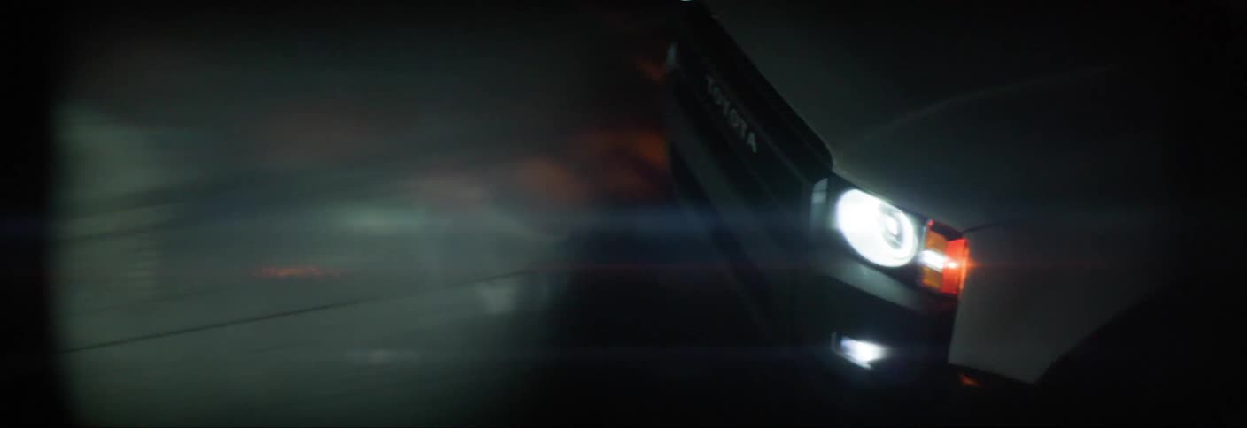 A dark car driving on a street at night with its hedslights illuminating the road
