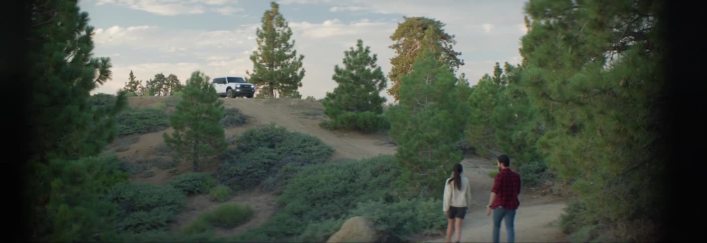 A man and a woman walking along a trail surrounded by trees with an offroad vehicle in the background