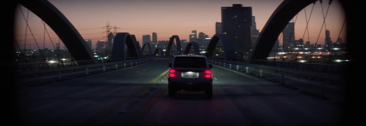 White car traveling across a bridge with a metropolis in the background