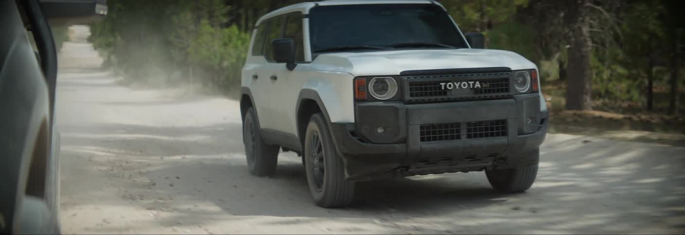 A white toyota suv drives through a dusty forest road kicking up a cloud of dust as it navigates the narrow path
