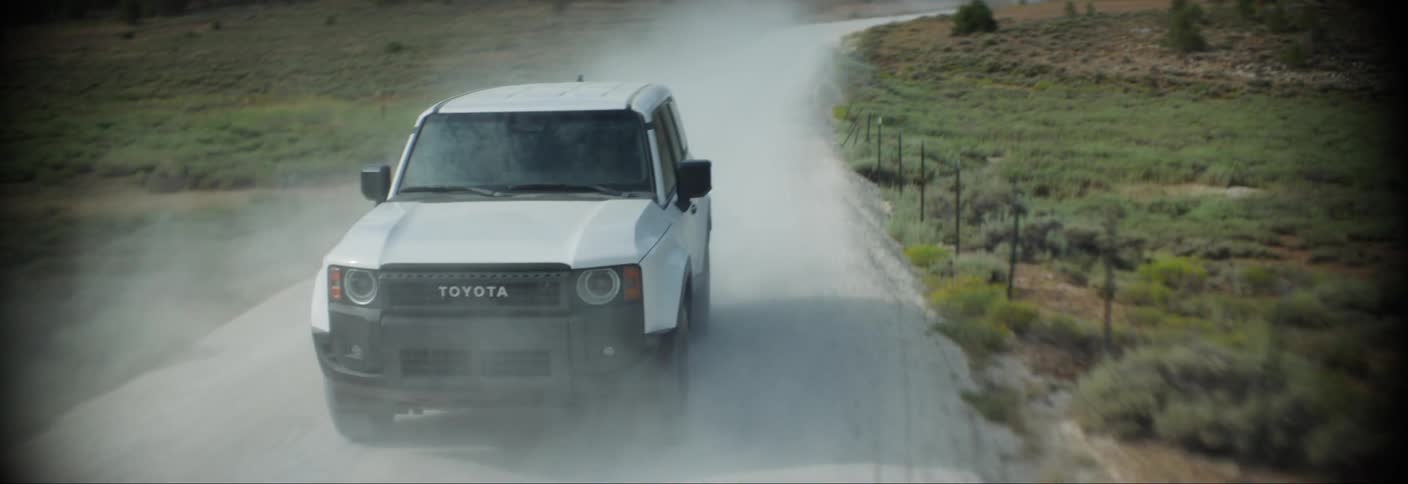 A white toyota land cruiser driving on a dirt road through a forested area kicking up a cloud of dust