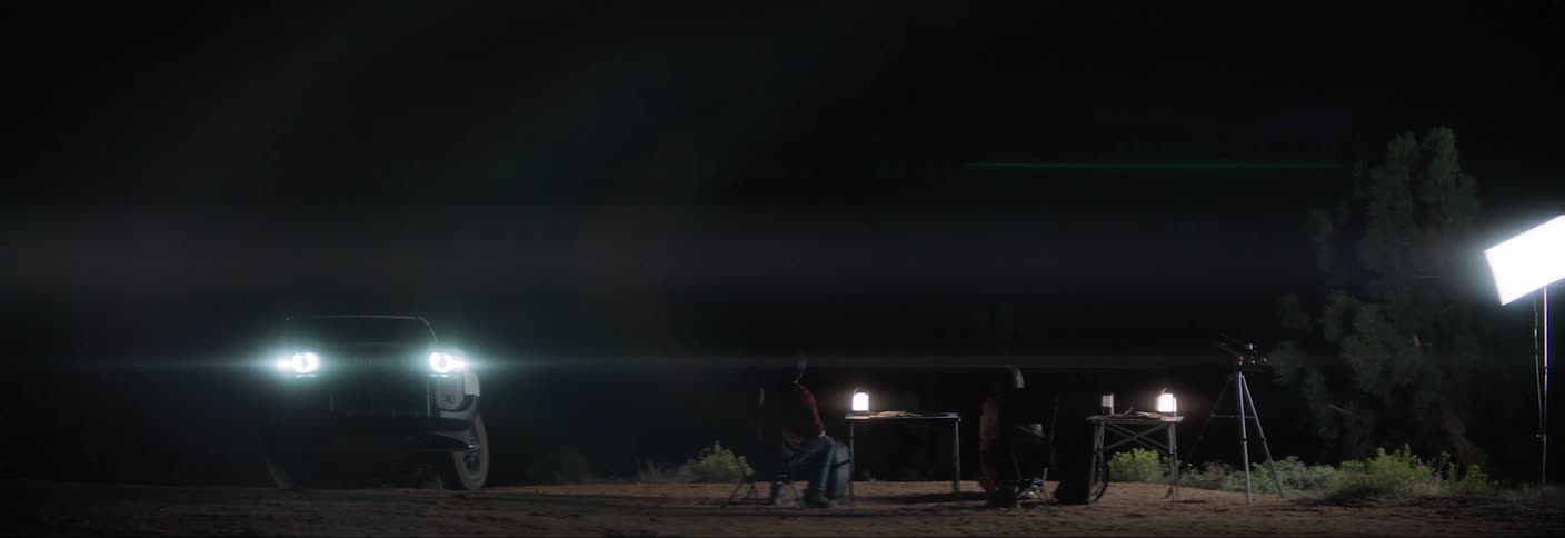 Overnight camp in an open area lit by headlights of a vehicle on the left and two people seated