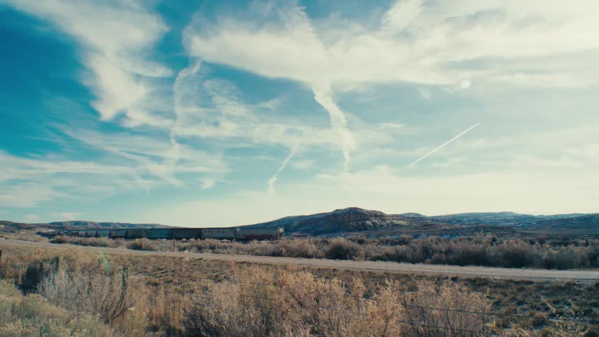 A long freight train winding through a vast arid landscape under a dramatic sky with fluffy white clouds and contrails stretching across the horizon