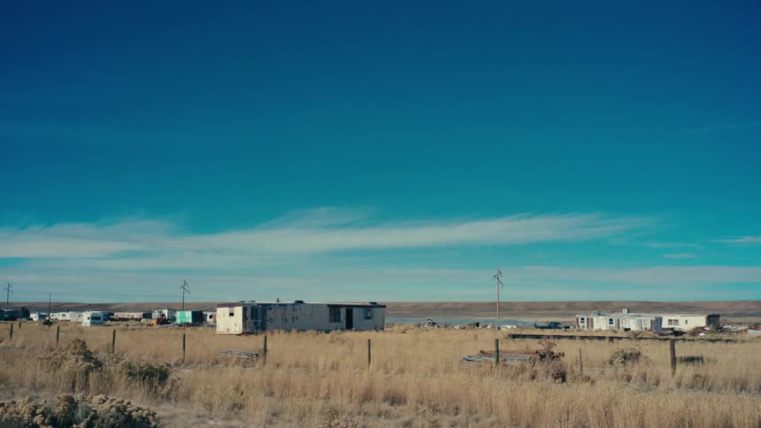 A cluster of weathered trailers and mobile homes sits abandoned in a vast desolate landscape surrounded by dry brown grass and a clear blue sky