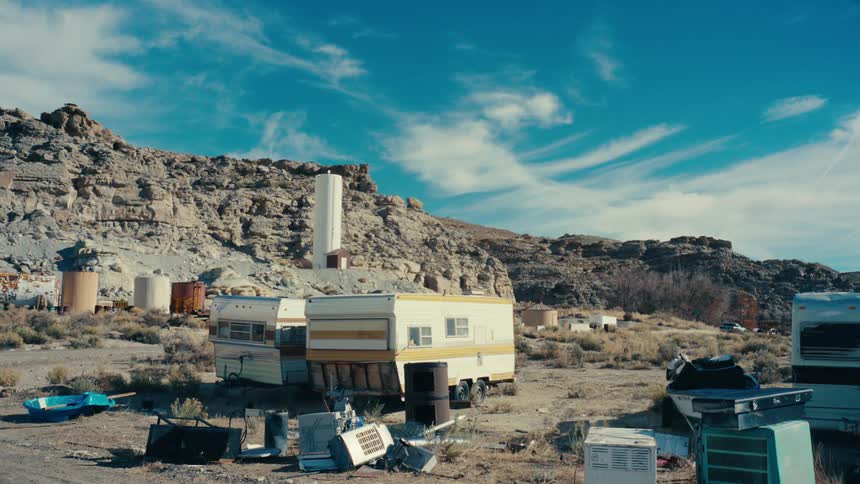 A desolate desert landscape with several old trailers scattered debris and industrial tanks near rocky hills and a clear blue sky
