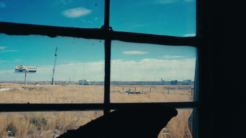 A view of an interior room with a silhouette of a dark window with a view of the outside environment where there is beige grass with a road where trucks are passing by