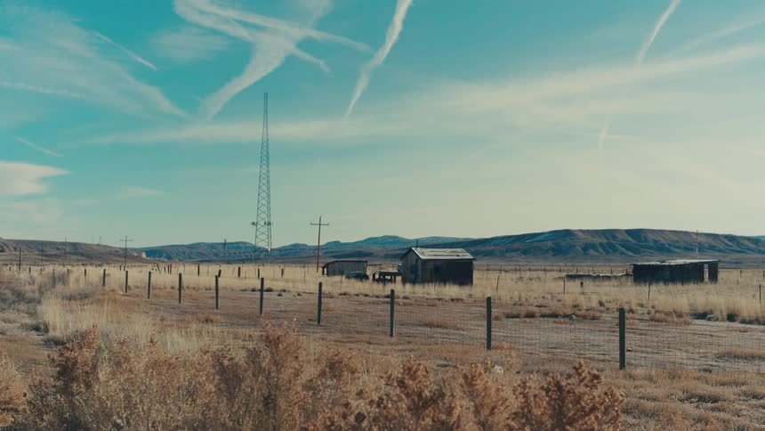 A static wide shot of a rural landscape with a tall antenna and a few small weathered buildings in the middle ground backed by low hills and a light blue sky with faint cloud streaks