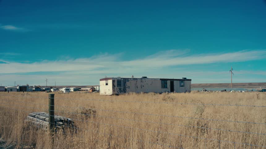 A weathered trailer sits abandoned in a vast open field surrounded by a cluster of other dilapidated structures under a clear blue sky