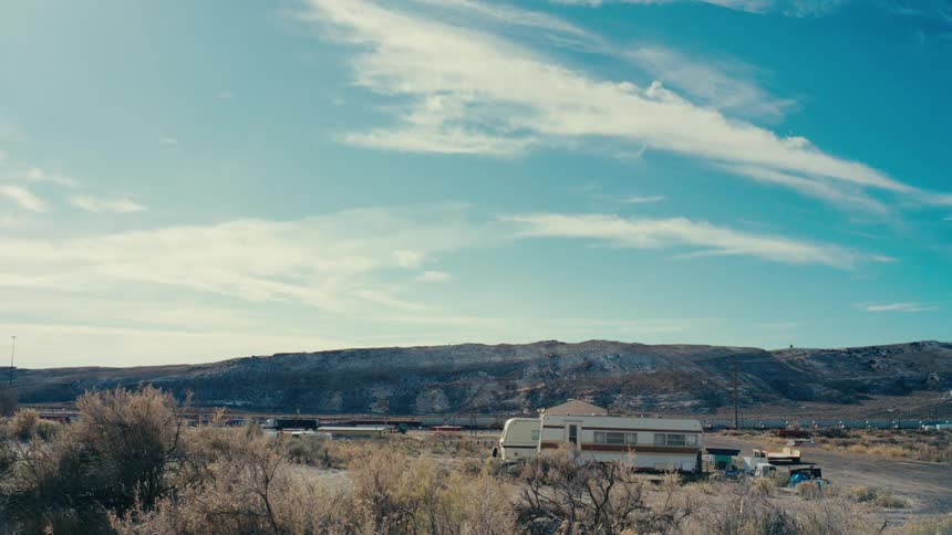 A cluster of weathered rvs and trailers sits abandoned in a desolate landscape surrounded by dry brown brush and a barren hill