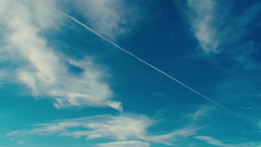 A wispy contrail streaks across a vast expanse of blue sky dotted with fluffy white clouds