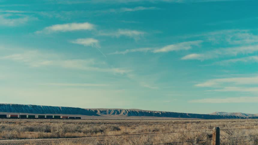A long freight train chugs across a vast open landscape dwarfed by the vast expanse of the horizon