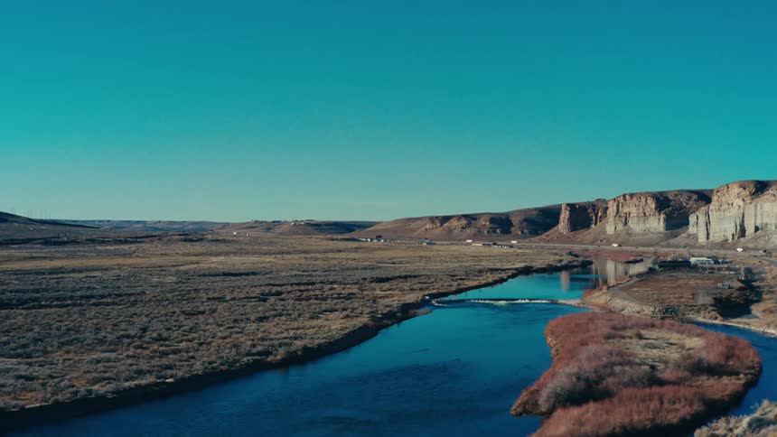 An aerial shot shows a wide clear blue river winding through a flat arid landscape under a cloudless sky with distant cliffs and a small settlement visible in the background