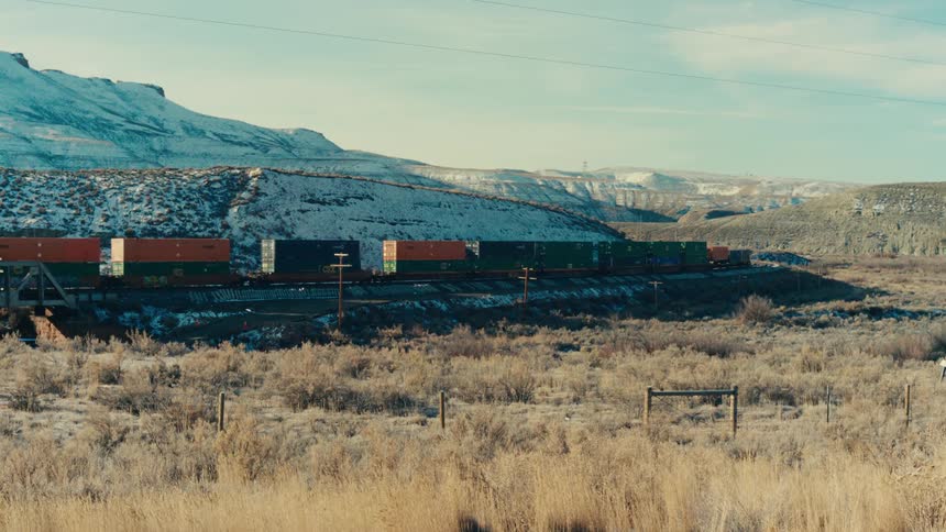 A long freight train chugs through a vast open landscape dwarfed by the snowcapped mountains in the distance