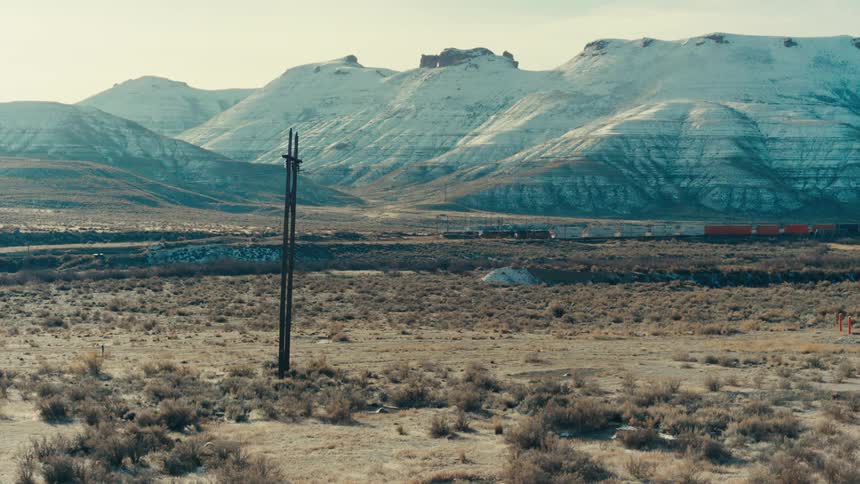 A long freight train chugs through a vast open landscape dwarfed by the snow capped mountains in the distance