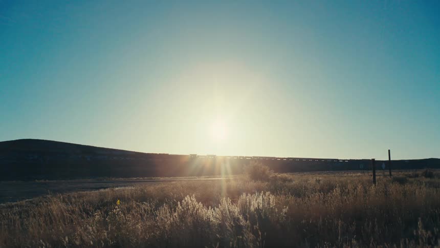 S a serene landscape with a sunlit field stretching towards a distant horizon framed by a clear blue sky and a gentle hill
