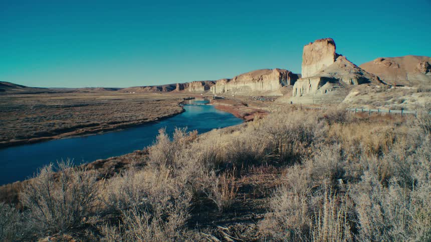 A winding river flows through a vast arid landscape framed by towering cliffs under a clear blue sky