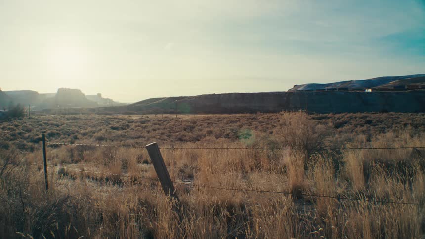 A vast open landscape with a fence running through a field of dry grass under a clear blue sky with the sun shining brightly in the distance a train chugs along the horizon