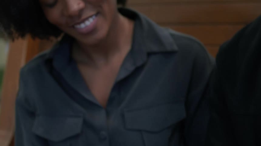 A happy woman with curly hair and a black shirt uses her smartphone while sitting on a bench next to a man