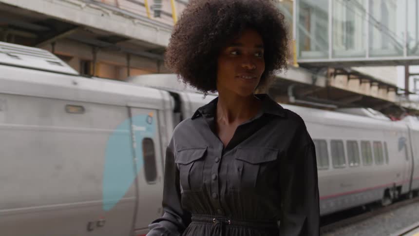 A woman wearing a black outfit stands on a train platform as a train passes by in the background