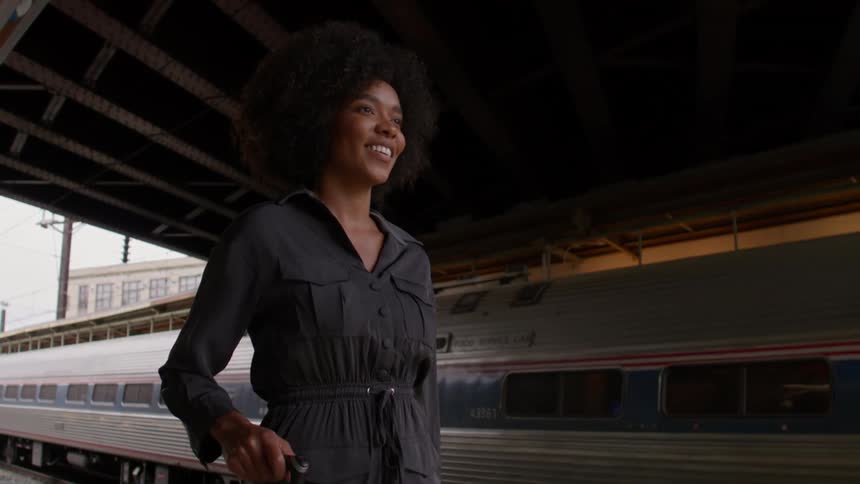 A woman with an afro wearing a dark coat holding a suitcase handle smiles as she walks on a train platform