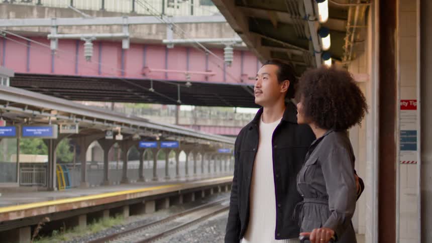 Man stands with his arm around a woman while they wait for the train with smiles