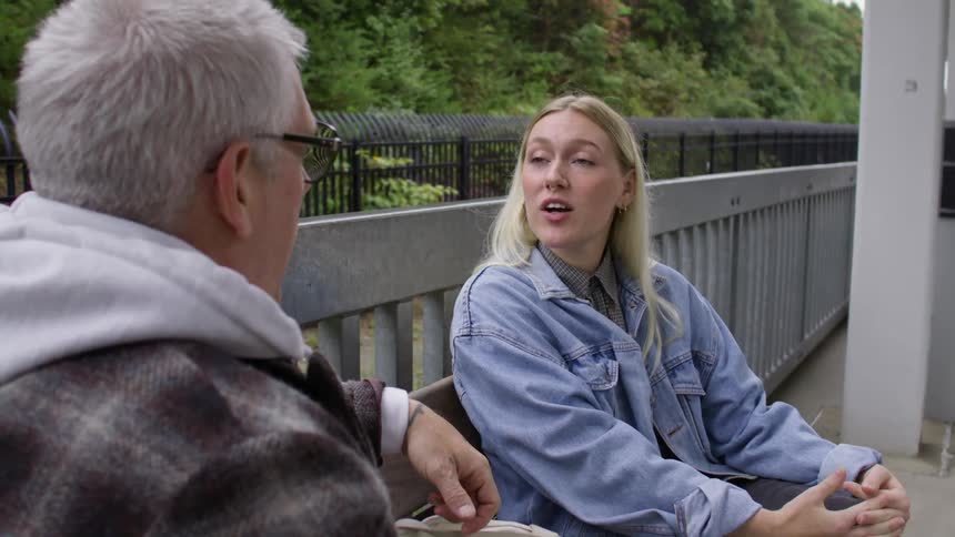 Blonde woman and a man sitting on a bench outdoors talking at a station