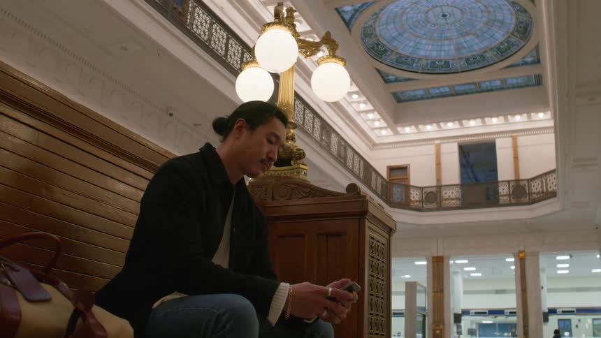 A man sits on a bench inside an ornate train station using his smartphone