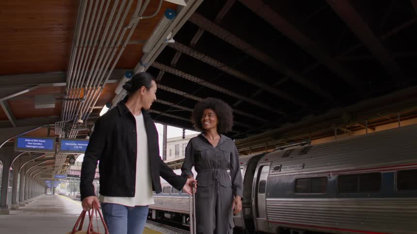 Two people walk hand in hand on a train platform with a train visible on the right side of the image