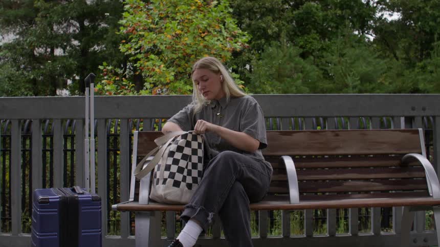 Blonde woman sitting on a bench outdoors opening her bag and taking out a smartphone