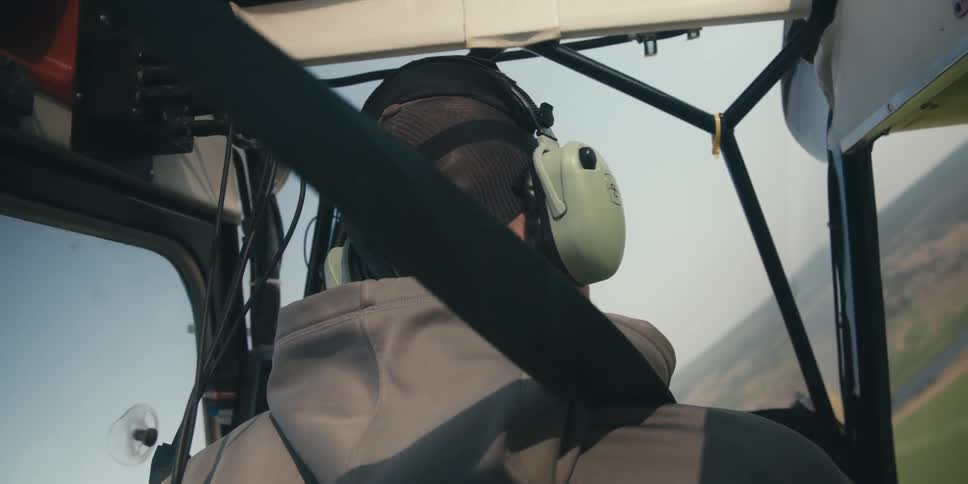 A person wearing a gray hooded sweatshirt a baseball cap and a headset sitting in the cockpit of an aircraft with a seatbelt in the foreground