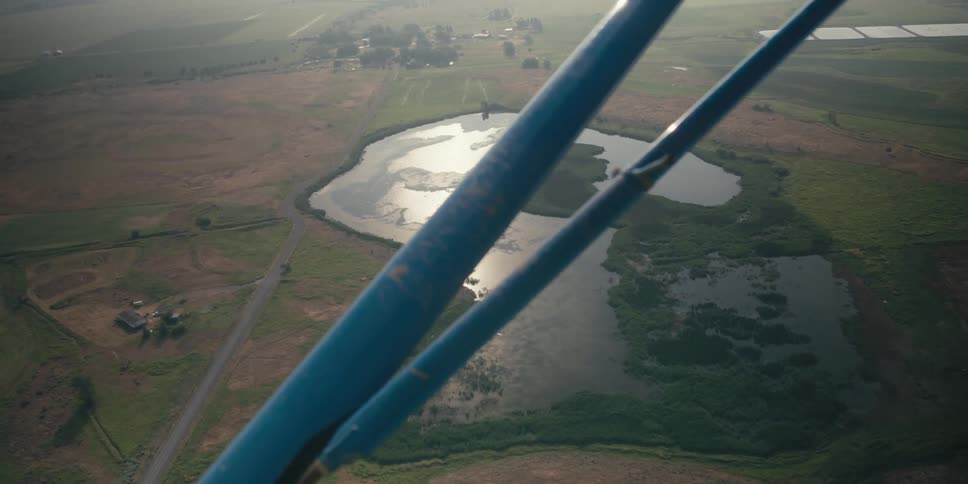 A small lake surrounded by green fields is visible from the cockpit of a small plane