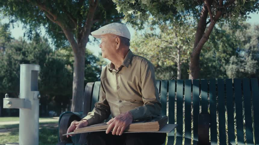Man smiles and looks around as he sits on a park bench with a photo album in his lap