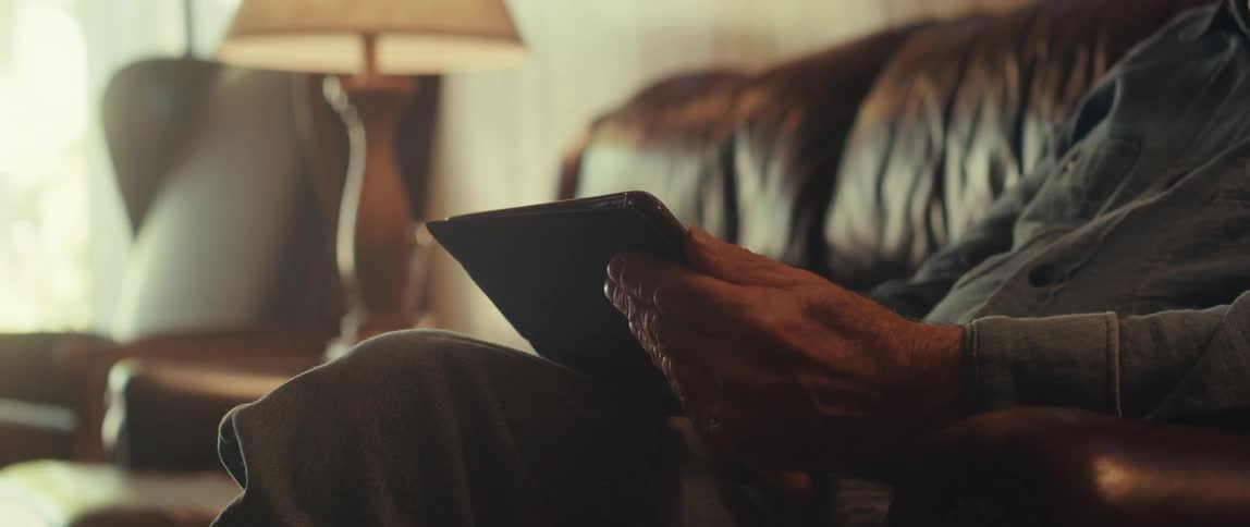 An elderly man sits on his sofa and scrolls on a tablet