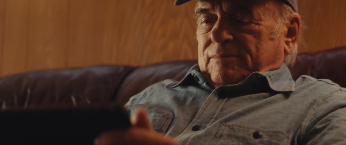 An elderly man in a cap is sitting on a sofa and looking at a tablet in his hand
