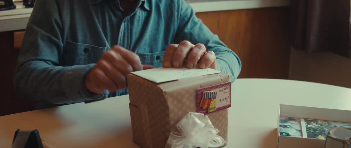 Man in a denim shirt is sitting at the table carefully viewing a gift wrapped with decorated paper and a white ribbon