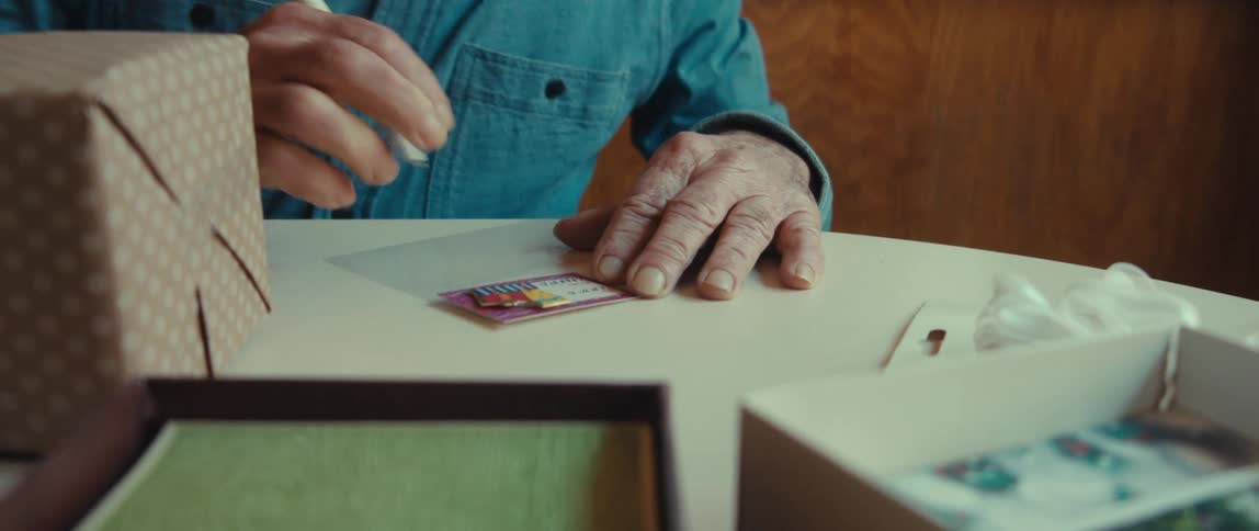 An elderly man with wrinkled hands writes on a small card and attaches it to a wrapped gift smiling as he works