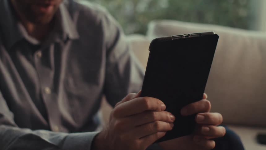 A person wearing a gray buttonup shirt is sitting on a couch having a video call on a tablet