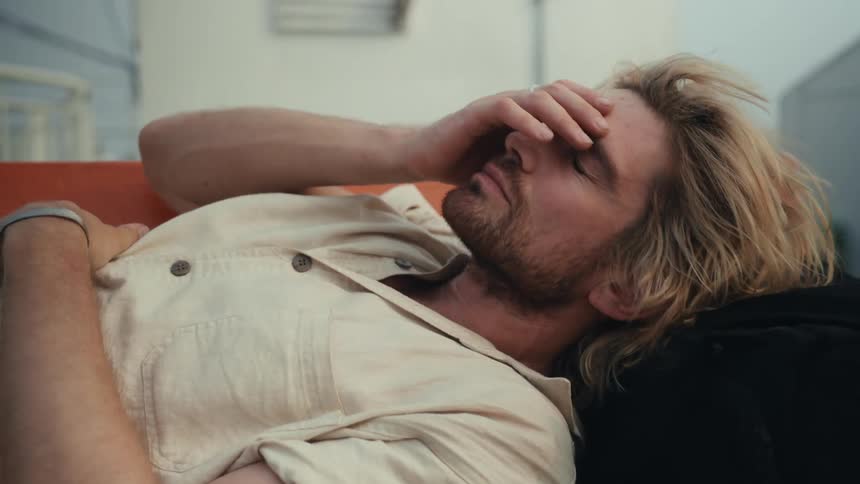 A blond man with long hair wakes up on a park bench during the day