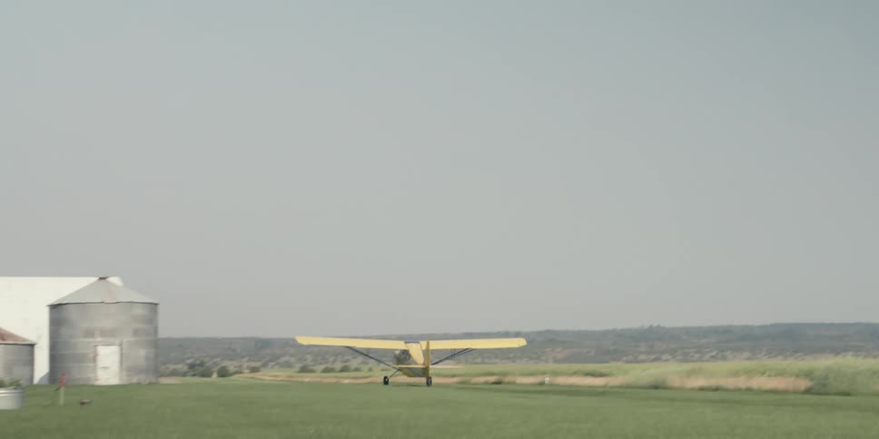 A small yellow airplane takes off from a grassy runway near two large metal silos and a white barn