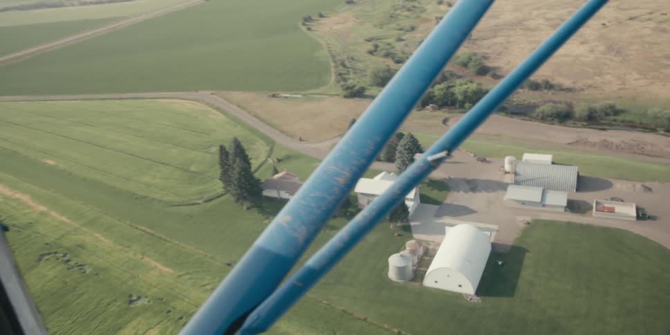 A pilot flying over a farmhouse surrounded by fields of grain