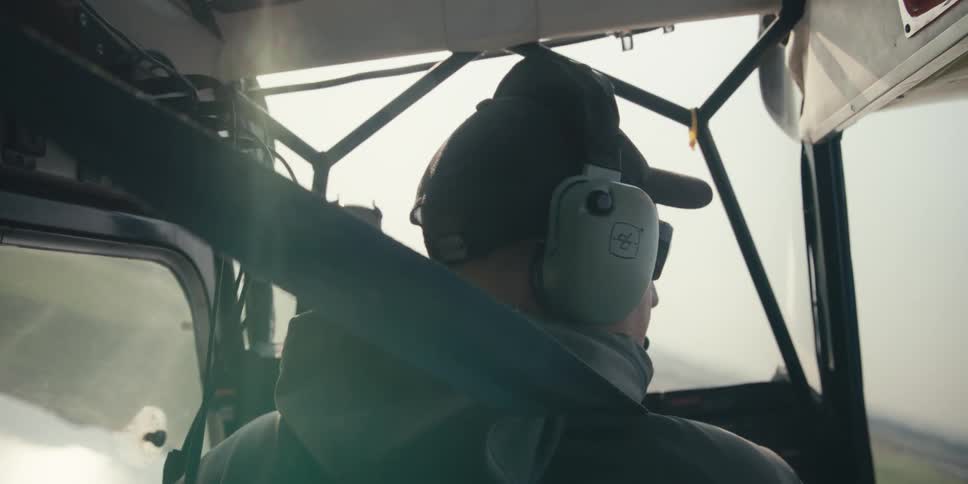 A person wearing a headset sits in the cockpit of an aircraft flying over a landscape with fields visible through the windows