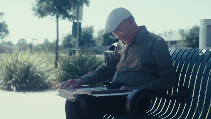 A man wearing a beret is sitting on a public bench and reading a large book while laughing