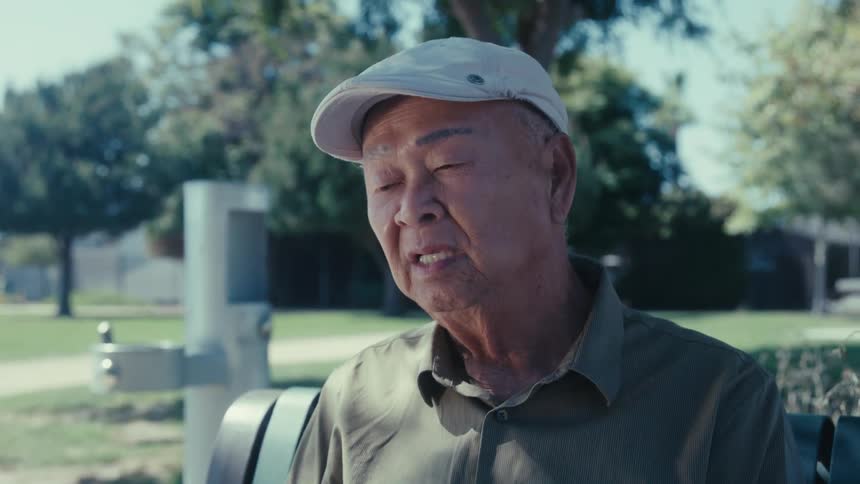 An elderly asian man sits on a park bench talking