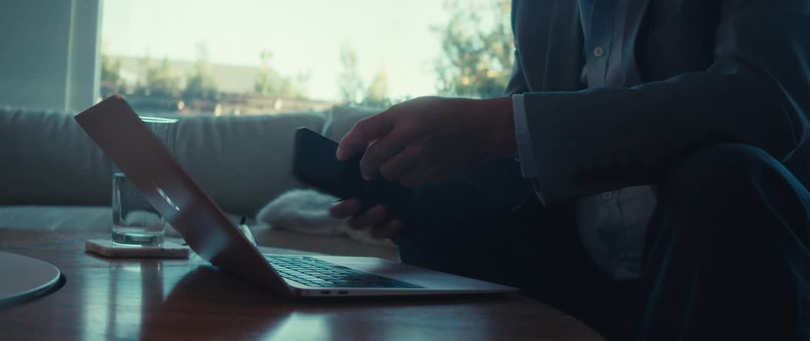 A man is using his cell phone and laptop while sitting on a couch