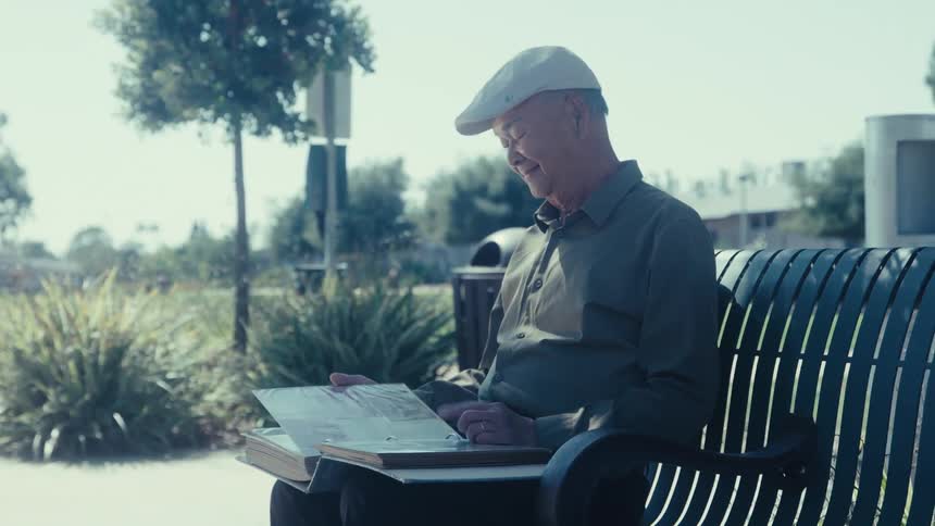 An elderly man wearing a cap and a longsleeved shirt is sitting on a park bench holding an open photo album