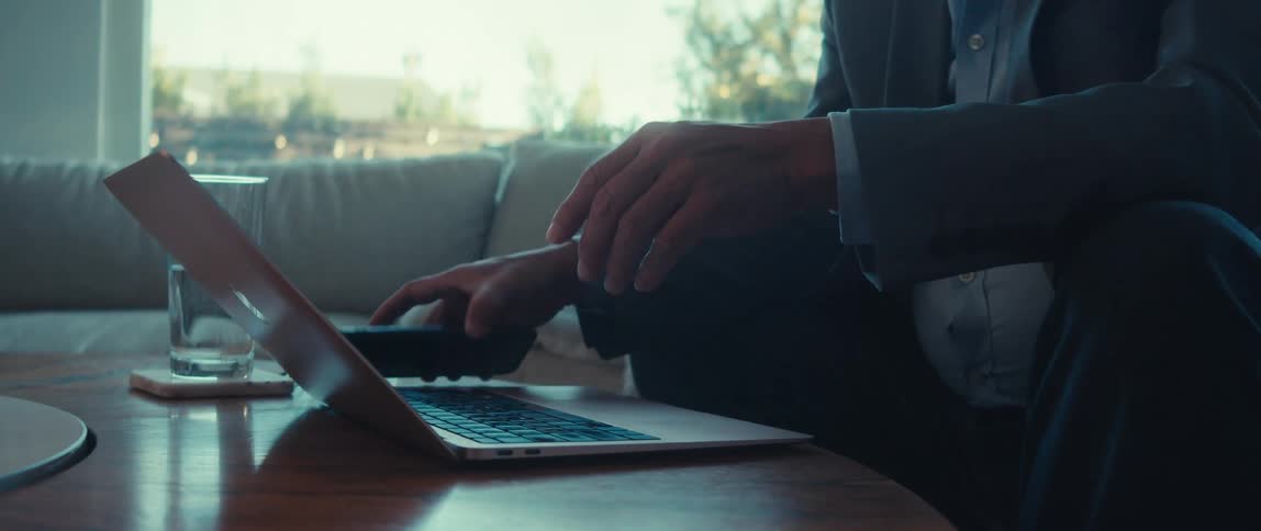 A man in a business suit sits on a couch at home working on a laptop before picking up a smartphone from the table