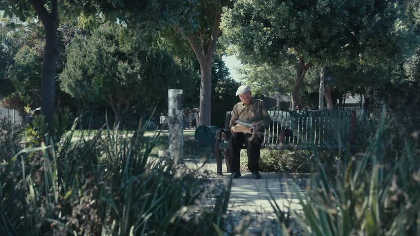 An elderly man walks toward a park bench sits down and opens a book to read