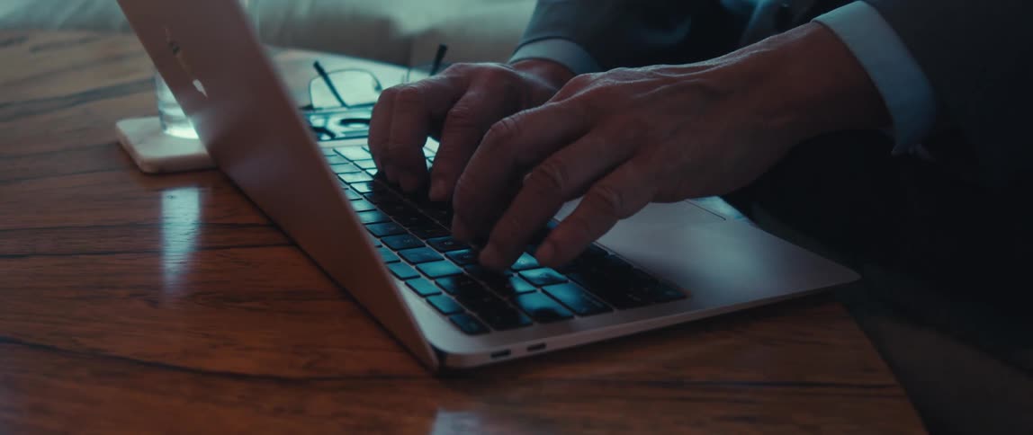A person is typing on a laptop keyboard placed on a wooden table with a pair of glasses and a glass of water nearby