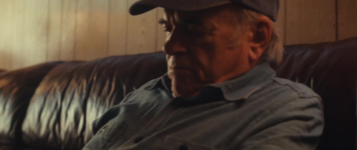 An elderly man in a cap and blue shirt sits on a leather couch looking at a tablet