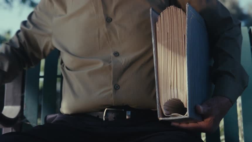 A person sitting on a bench has a book in his hand and puts it on his arm and gets up and leaves